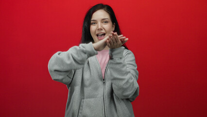 Woman smiling in gray hoodie making money gesture against red background showing playful confidence...