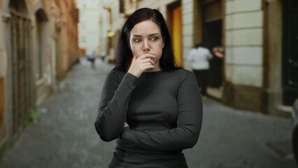 Woman with brunette hair standing in an outdoor street setting, crossing her arms and expressing...