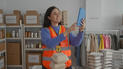 Woman videoconferencing while volunteering indoors in donation center surrounded by supplies...