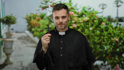 Priest holding cross outdoors in garden setting with thoughtful expression and clerical attire,...
