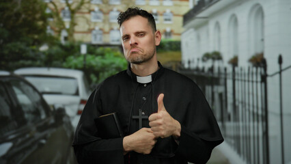 Priest in black robe with bible and cross gives thumbs up on urban street with blurred buildings...