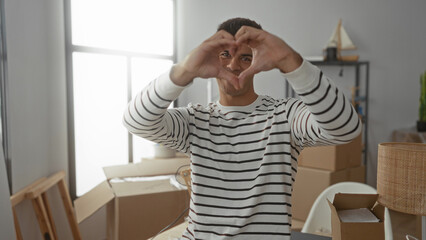 Young man smiling indoors forming heart shape with hands surrounded by moving boxes in new home hinting at happiness and fresh start in apartment environment.