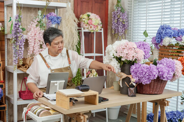 Asian senior florist checking laptop screen while receiving flower order online arranging hydrangea...