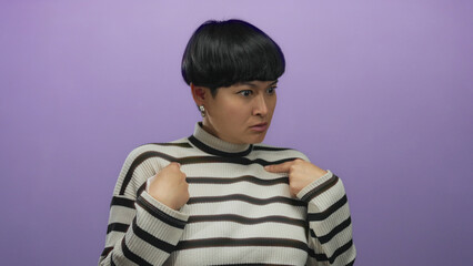 Woman pointing to herself against an isolated purple background showing surprise wearing a striped sweater with bold earrings and a short hairstyle emphasizing expressive reactions