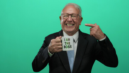 Senior man in a business suit smiles while holding a mug with 'i am the boss' text, pointing to his...