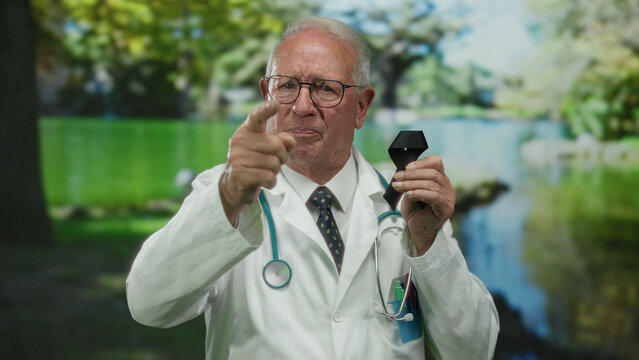 Senior male doctor in white coat standing outdoors in a park holding a black ribbon while gesturing with his hand, surrounded by lush green scenery.
