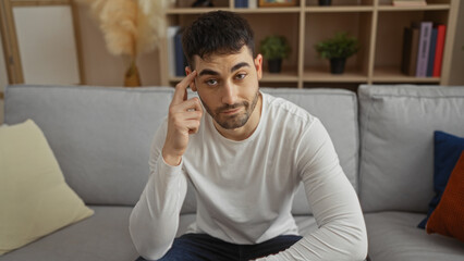 Young hispanic man making a think gesture while sitting in a cozy living room at home, wearing a white shirt with a thoughtful expression.
