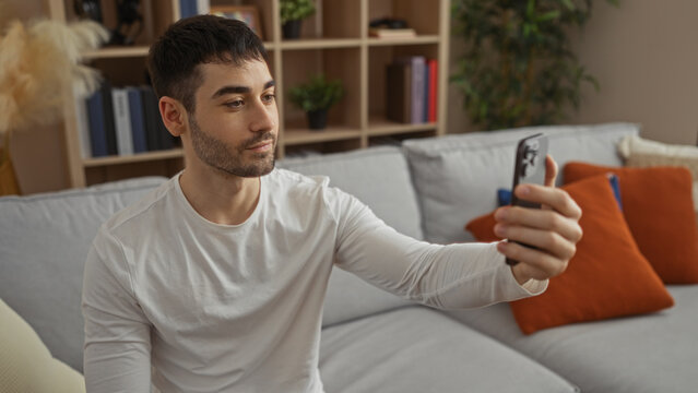 Young hispanic man taking a selfie in a cozy living room interior, capturing a relaxed moment at home with modern decor and soft lighting for a personal portrait.