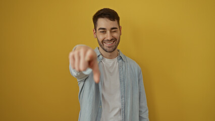 Young man winking and pointing at the camera over a bright yellow background, capturing a playful...