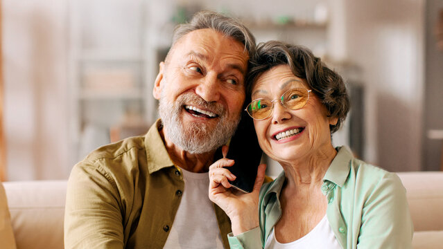 Happy senior husband and wife talking with relatives, holding cellphone between them, listening and smiling