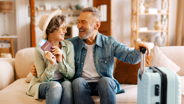 Joyful retired senior couple ready for trip, sitting on sofa at home, looking and smiling at each other, waiting for vacation