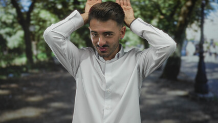 Man in white shirt with ear stud raises hands above head amid green trees in street with pursed...