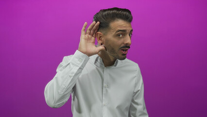 Young hispanic man cups hand to ear in studio with purple background; listening attention curiosity.