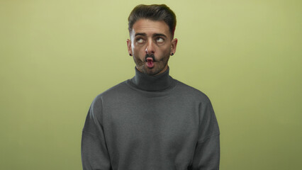 Obraz premium Young hispanic man purses lips and glances sideways wearing grey turtleneck sweater in olive green studio; curiosity.