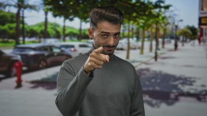 Man wearing turtleneck sweater points finger forward on city street lined with parked cars;...