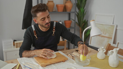 Young hispanic man shaping white clay with bare hands on wooden board in studio; concentration...