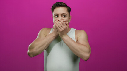 Young man with beard covers mouth with hands, standing against vibrant pink wall in isolated...