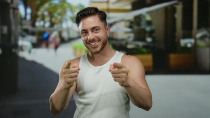 Young man with beard smiling and pointing on a city street terrace in an outdoor urban coffee...
