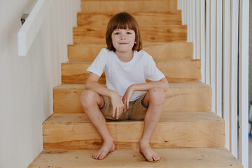 Happy boy sitting on wooden stairs, wearing a white t-shirt and shorts. This image captures a carefree moment of childhood, embodying joy and innocence. © SHOTPRIME STUDIO