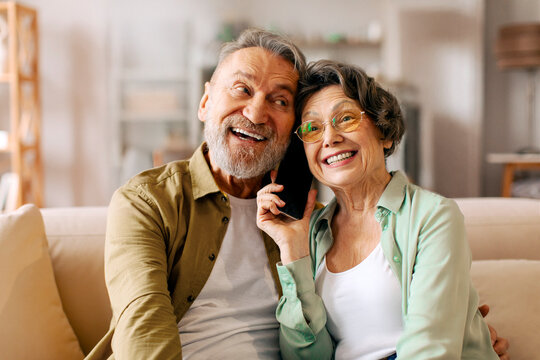 Happy senior husband and wife talking with relatives, holding cellphone between them, listening and smiling - Powered by Adobe