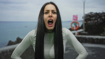 Woman with long hair stands at seaside promenade looking surprised with ocean in the background under cloudy sky, showcasing expressive emotions and outdoor scenery.