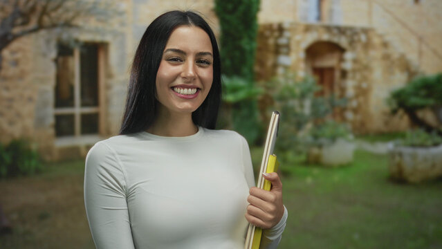 Woman smiling outdoors pointing and holding books near a rustic building with stone walls and lush greenery in the background capturing a cheerful academic scene.