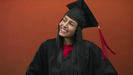 Young hispanic woman smiling in graduation uniform with cap in front of orange background,...