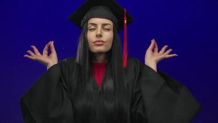 Woman meditating in graduation cap and gown, eyes closed against blue background, embodying calm...