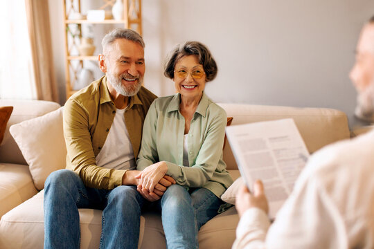 Happy European senior spouses holding hands while having consultation with real estate agent or psychologist, sitting on sofa. Specialist's service offer