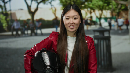 Woman with long hair in red jacket holds helmet while standing in urban street with trees and...