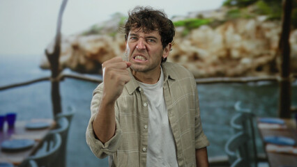 Hispanic man expressing anger at a beachside venue with sea view in the background, illustrating a...