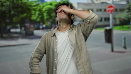 Fototapeta premium Man standing outdoors on a street with trees and buildings in the background covering his face with his hand.