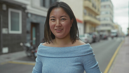 Fototapeta premium Young woman smiling on city street with modern buildings around in an urban setting, captured outdoors showcasing casual clothing in a vibrant metropolitan atmosphere.