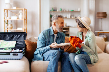 Excited senior couple packing clothes for trip preparing for vacation, sitting on sofa in living...