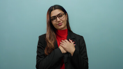 Young hispanic woman in glasses and a black blazer with hands on heart expressing gratitude against...