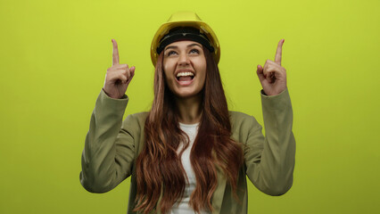 Smiling young hispanic woman architect in hard hat points upward against isolated yellow...