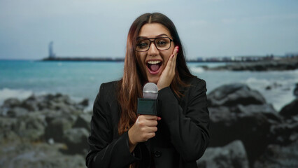 Woman holding microphone on beach as reporter on outdoor seaside location wearing glasses appears...