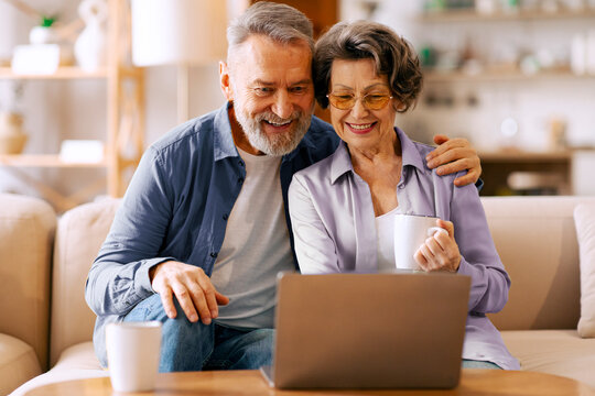 Positive senior spouses using laptop and drinking coffee, couple browsing internet, sitting on sofa at home. Gadgets and leisure for elderly people