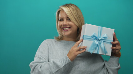 Young blonde woman holds gift box with blue ribbon by her face against teal background in studio;...