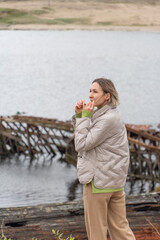 Woman in a beige puffer jacket and sunglasses standing near rusted shipwrecks by the shore, looking...