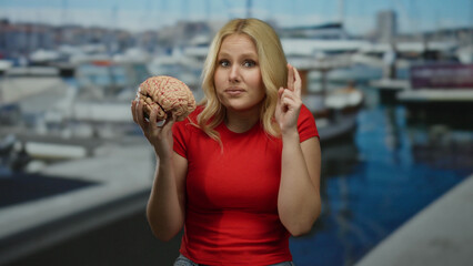 Fototapeta premium Woman holding a brain model with fingers crossed in a marina backdrop featuring boats and ships, embodying hope and intellect.