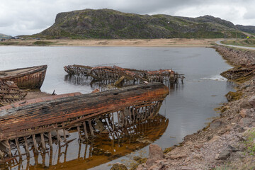 Rusted wooden and metal shipwrecks partially submerged in shallow coastal waters against a backdrop...
