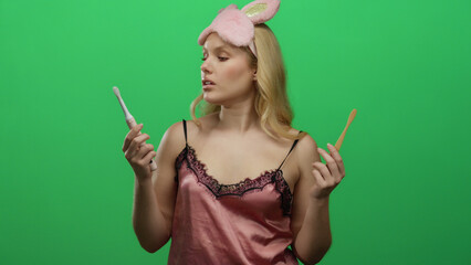 Blonde woman in pink nightwear holding a toothbrush against a green background wearing a pink sleep...