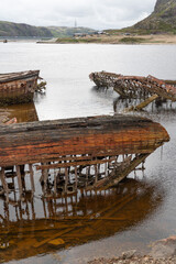 Rusted wooden and metal shipwrecks partially submerged in shallow coastal waters against a backdrop...