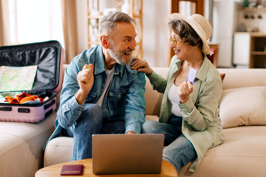 Happy senior spouses receiving mail or booking hotel online, clenching fists while packing clothes for travel, sitting with suitcase on couch - Powered by Adobe
