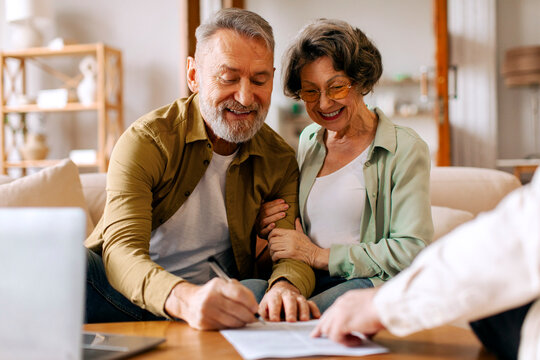 Happy retired senior couple signing real estate lease contract while meeting with realtor, elderly man and woman filling paper form