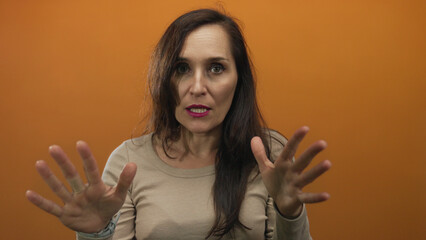 Young hispanic woman raises bare hands palm outward in calm gesture in front of orange studio wall;...