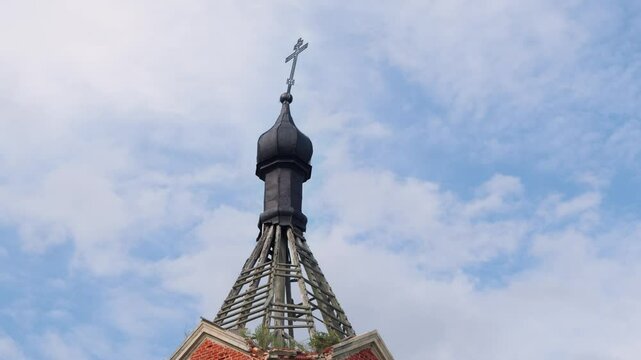 A close-up of a small black Orthodox dome with a lopsided cross on top of a brick-and-wooden abandoned bell tower against background of blue sky with clouds.