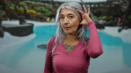 Senior woman with grey hair listens intently by a pool in a hotel resort setting, exhibiting...