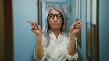 Senior woman with grey hair in hotel corridor signaling no with fingers and focused expression,...